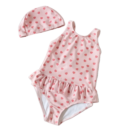 Flat lay view of a toddler girl's pink one-piece swimsuit featuring a red heart print and ruffle waist, shown on a white background with a matching swim cap.