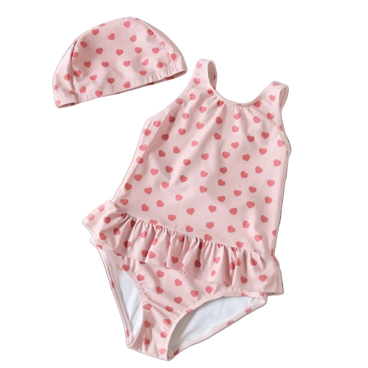 Flat lay view of a toddler girl's pink one-piece swimsuit featuring a red heart print and ruffle waist, shown on a white background with a matching swim cap.