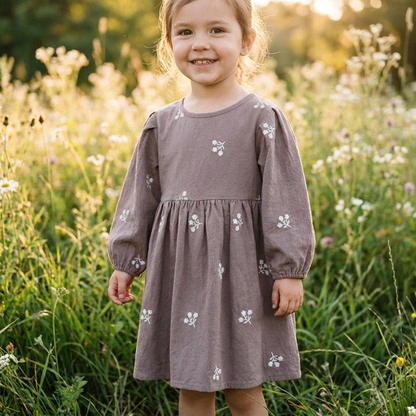 A little girl smiling in tall green grass wearing the gray long-sleeve cottagecore floral dress.