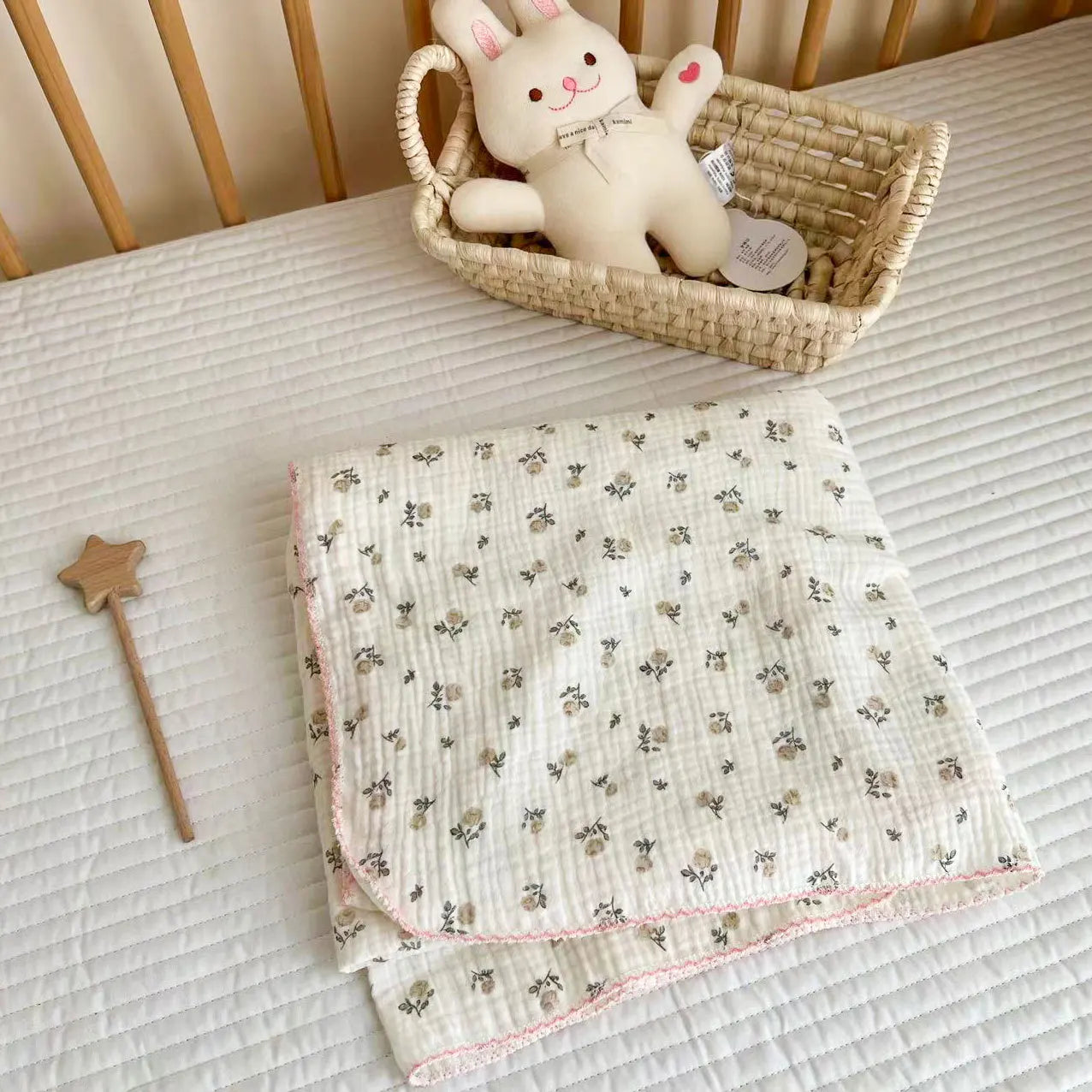 Folded gray and pink rose print cotton muslin swaddle blanket with a pink scalloped edge resting in a nursery crib.