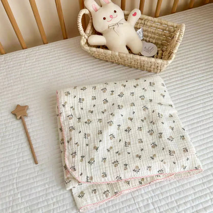 Folded gray and pink rose print cotton muslin swaddle blanket with a pink scalloped edge resting in a nursery crib.