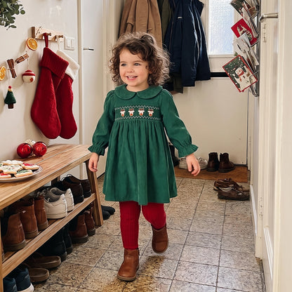 Young girl wearing a green long-sleeve smocked corduroy Christmas dress with red tights indoors.