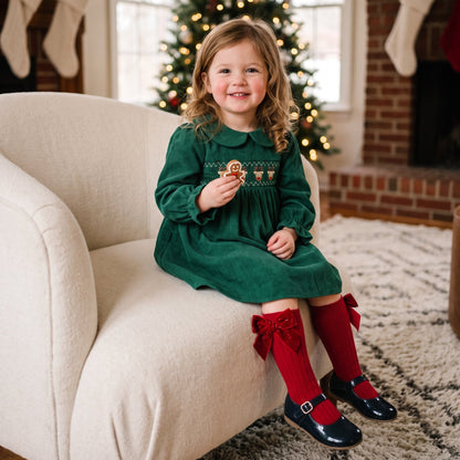 Toddler girl sitting on a chair wearing a green corduroy Christmas dress with red knee socks and holiday decor
