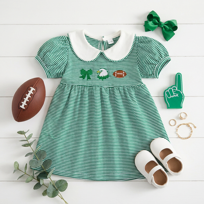 Flat lay of the green striped game day dress styled with white toddler shoes, a miniature football, and a green hair bow.