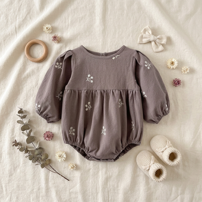 Flat lay of the cozy grey floral embroidered long-sleeve baby romper, beautifully styled with cream knit baby booties, a fabric hair bow, a wooden teething ring, and dried eucalyptus and flowers on a soft cream muslin background.
