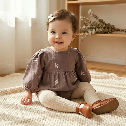 Smiling baby girl sitting indoors wearing the grey floral embroidered long-sleeve romper with cream tights.