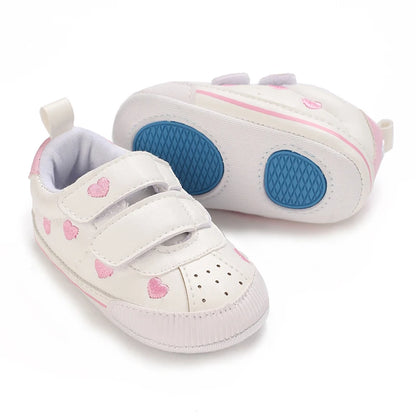 Pair of white heart print baby sneakers with one shoe turned over to reveal the soft sole with blue anti-slip grip pads.