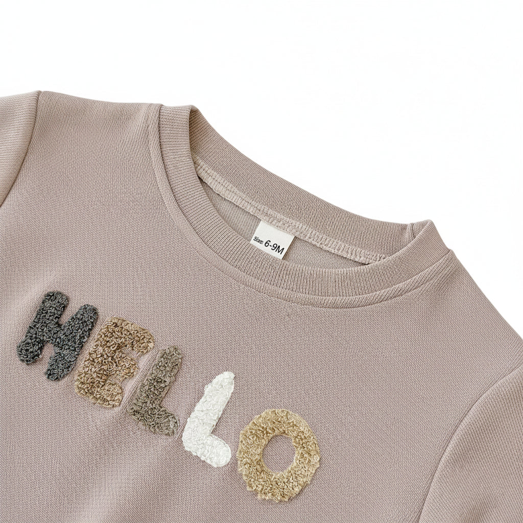 Close-up of the textured towel letter embroidery spelling HELLO on the chest of the beige baby sweatshirt.