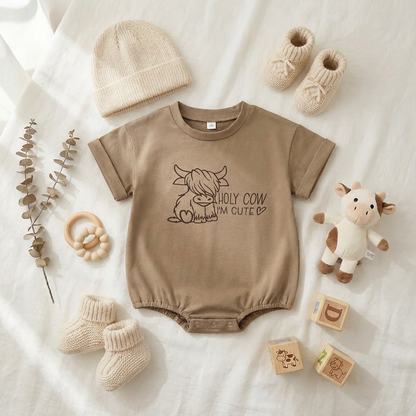 Styled flat lay of the light brown Highland cow baby romper surrounded by a beanie, booties, a small cow plush toy, and wooden blocks.