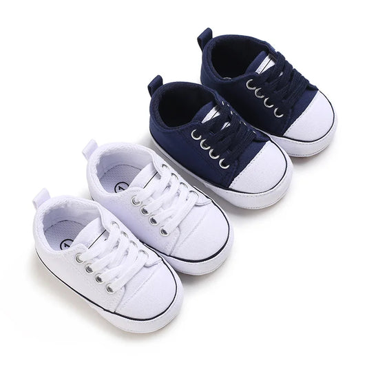 Two pairs of classic infant athletic canvas sneakers in white and navy blue, featuring soft soles, white toe caps, and adjustable laces for first walkers.