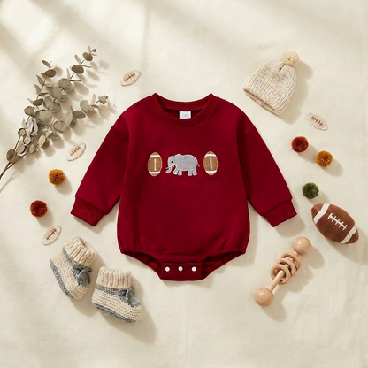 Styled flat lay of a crimson red game day infant bubble romper with elephant embroidery paired with baby shoes, a hat, and wooden toys.