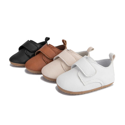 Four pairs of infant first walker faux leather shoes lined up in black, brown, beige, and white.