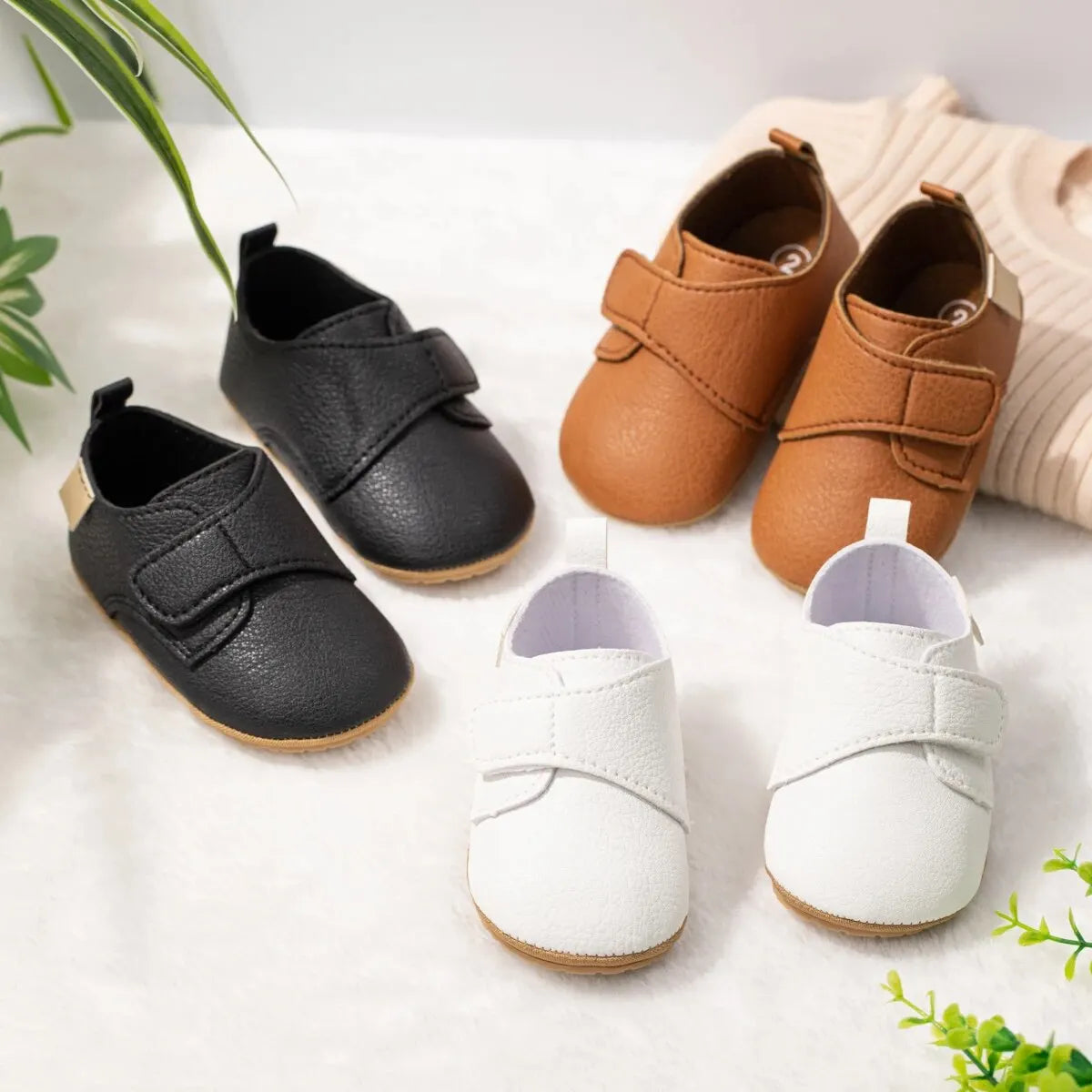 Top-down view of infant first walker faux leather shoes in black, brown, white, and beige scattered playfully on a white surface.