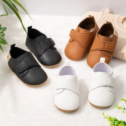 Top-down view of infant first walker faux leather shoes in black, brown, white, and beige scattered playfully on a white surface.