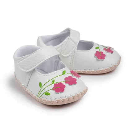 Angled view of white soft sole baby girl crib shoes featuring pink floral embroidery and adjustable hook-and-loop straps.