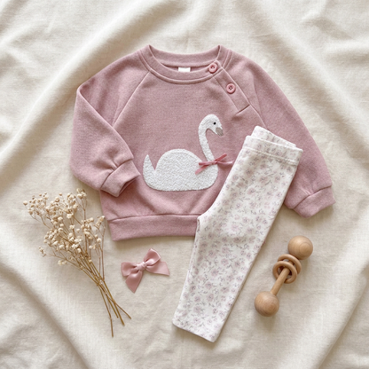 Flat lay aesthetic display of the infant girl swan embroidery sweatshirt and floral print trousers next to a small wooden rattle and hair bow on a neutral fabric background.