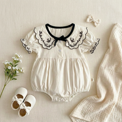 Flat lay of the ivory and black embroidered bubble romper styled with baby shoes and a bow for a complete summer outfit.
