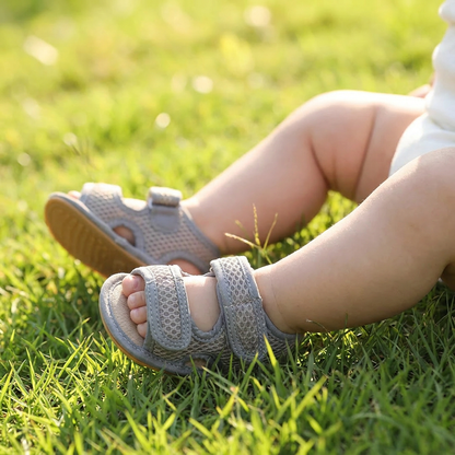 Close-up of a baby wearing grey first walker sandals in the grass, highlighting the flexible sole for outdoor play.