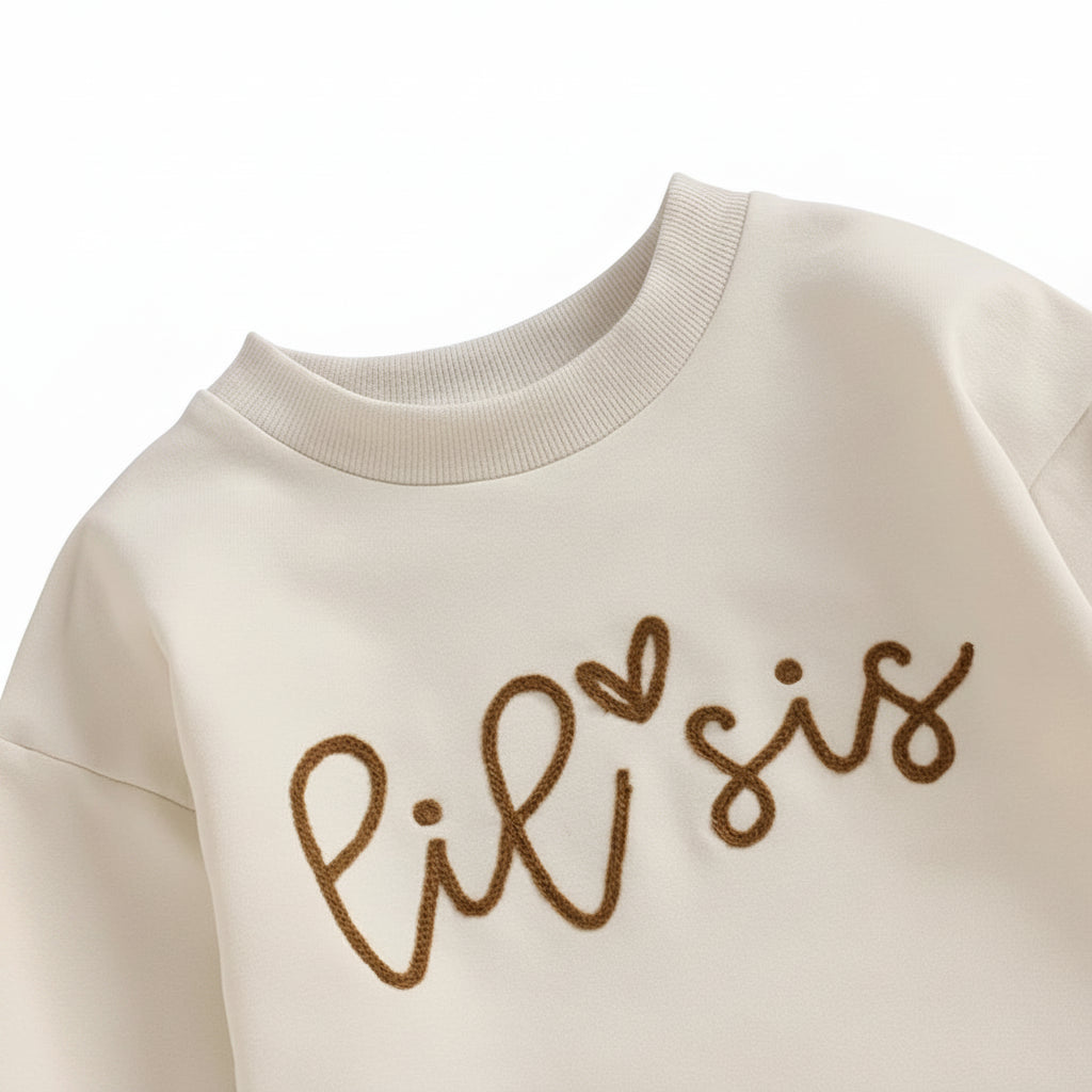 Detailed close-up view of the fine brown script lettering reading Lil Sis on the chest of the cream infant romper.