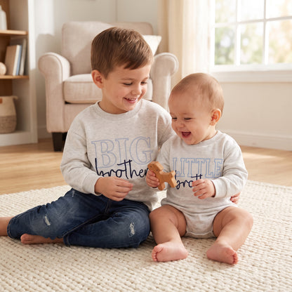 A toddler boy and a baby boy smiling and sitting together on a rug, wearing matching grey Big Brother and Little Brother long-sleeve outfits.
