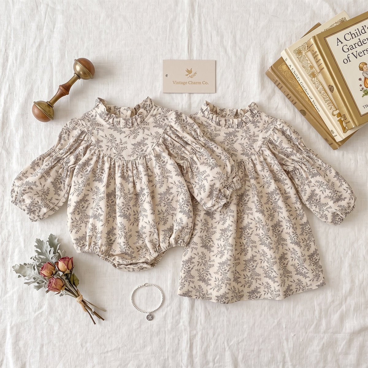 Flat lay showcasing a matching sister outfit set, featuring a cream and gray floral baby bubble romper and a girls long-sleeve dress styled with a vintage rattle, books, and dried flowers.