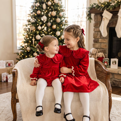 Matching sister cherry red velvet Christmas outfits featuring baby romper and toddler dress by Christmas tree