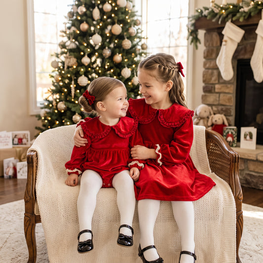Matching sister cherry red velvet Christmas outfits featuring baby romper and toddler dress by Christmas tree