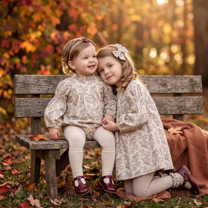 Two young sisters wearing matching vintage-inspired cream and gray botanical outfits—a baby bubble romper and a long-sleeve girls dress—posing together by a wooden bench for fall family photos.