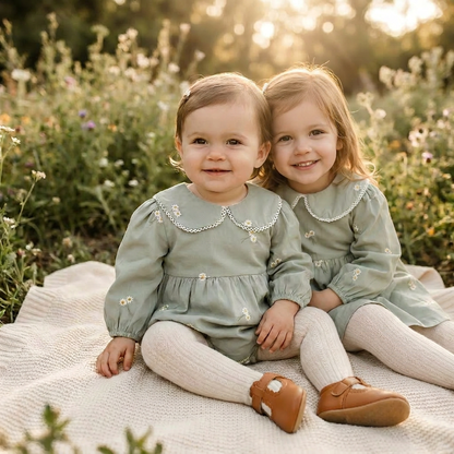 Two young sisters sitting together on a blanket in an outdoor field, wearing matching sage green embroidered outfits with cream tights and leather shoes.
