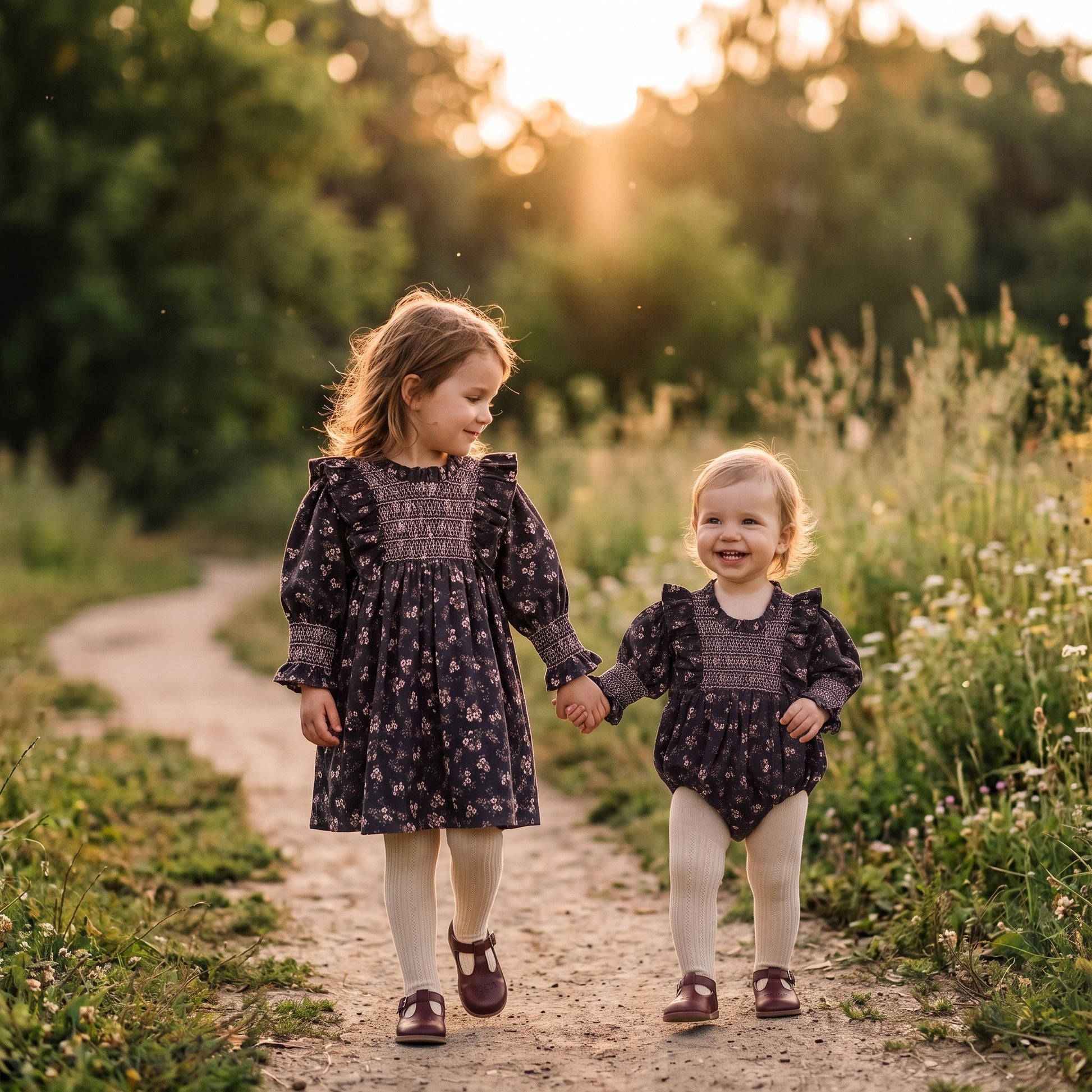 Little girl wearing matching grey floral smocked long sleeve dress walking hand in hand with baby sister in matching romper outdoors for fall family photos.