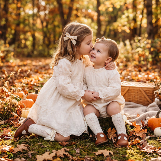 Two young sisters sitting outdoors in autumn wearing matching ivory floral embroidered dress and baby bubble romper.
