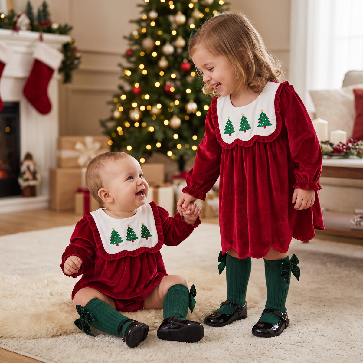 Baby and toddler sisters wearing matching red velvet Christmas tree outfits in front of a Christmas tree