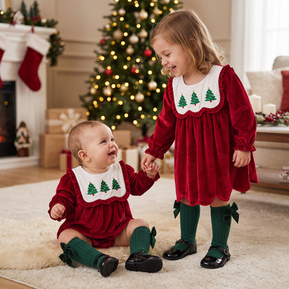 Baby and toddler sisters wearing matching red velvet Christmas tree outfits in front of a Christmas tree