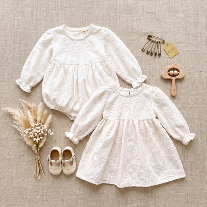 Flat lay arrangement of matching white floral embroidered toddler dress, baby bubble romper, and neutral nursery accessories.
