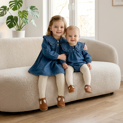 Two smiling sisters sitting on a cream couch wearing matching blue denim outfits, featuring a toddler dress and a baby romper styled with white tights and brown shoes.