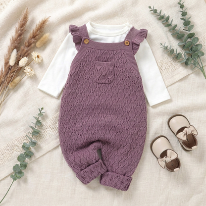 Styled fall baby outfit featuring mauve knit overalls layered over a white shirt, paired with baby shoes and greenery on a textured rug.