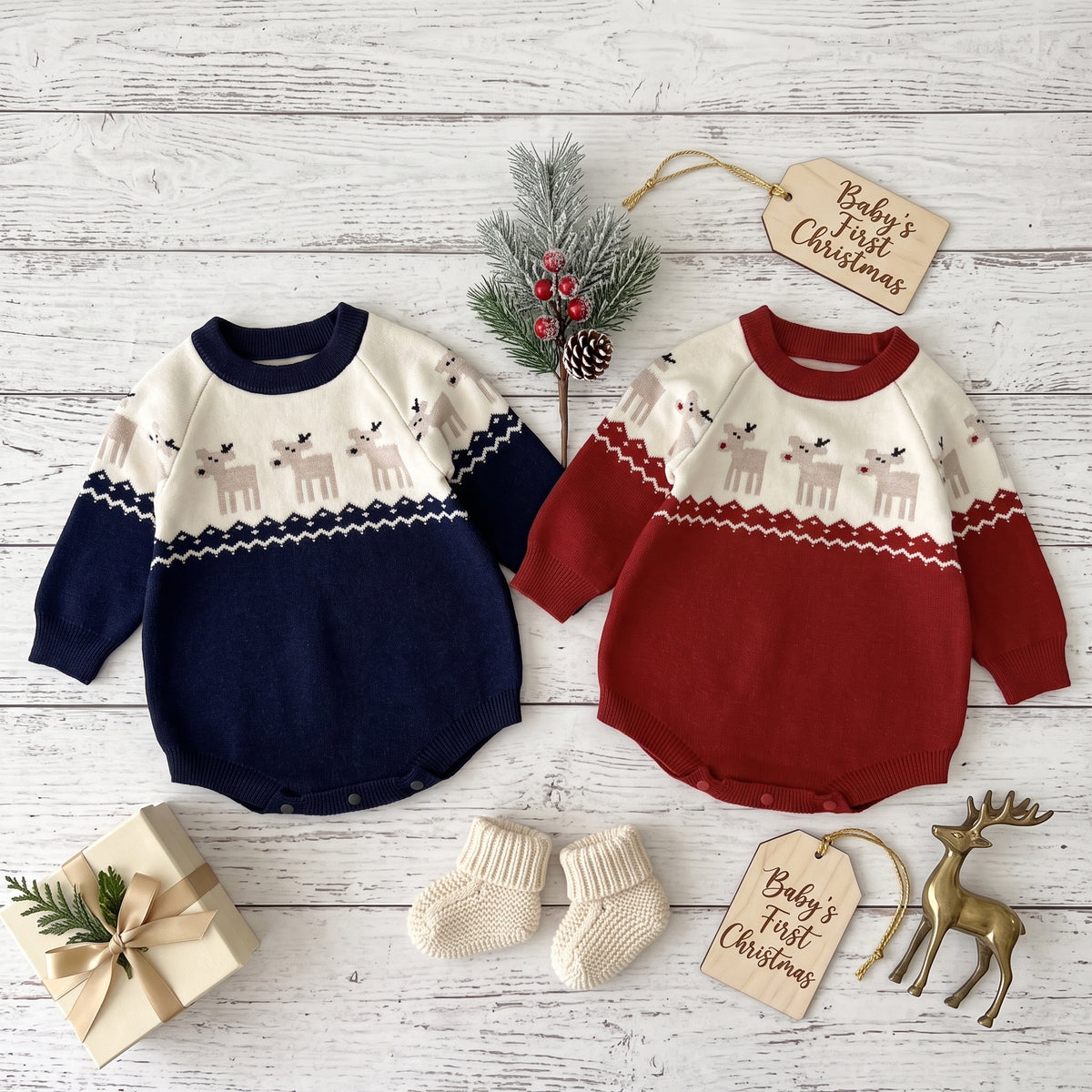 Navy and red baby Christmas sweater rompers laid flat with winter props, pine sprigs, and knit baby booties.