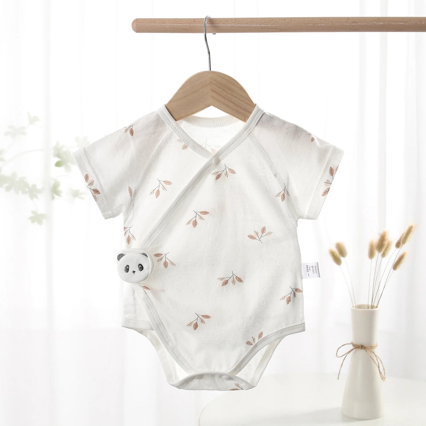 Earthy neutral beige botanical leaf print baby bodysuit hanging on a minimalist wooden clothes hanger against a soft-lit background curtain.