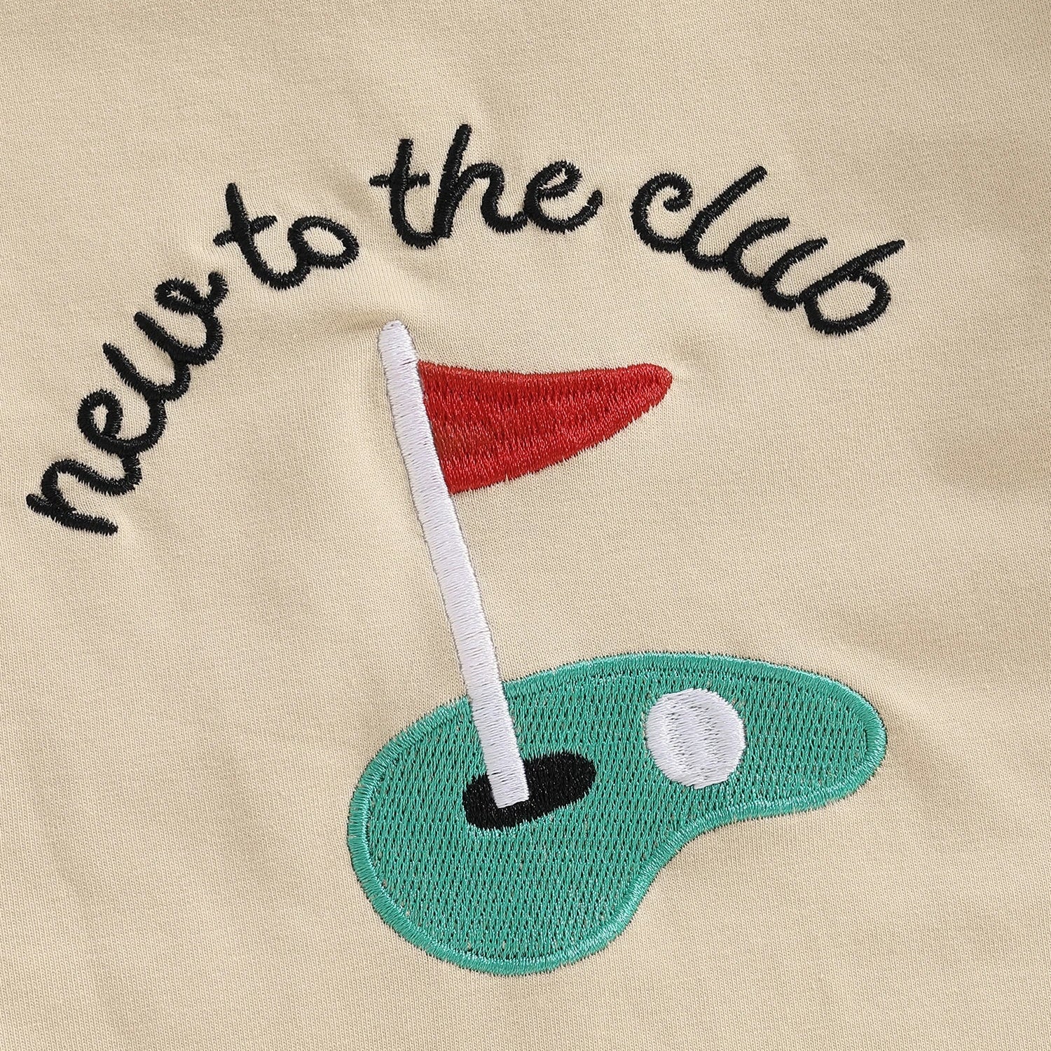 Close-up detail of the "new to the club" text and putting green embroidery on the beige long-sleeve baby romper.