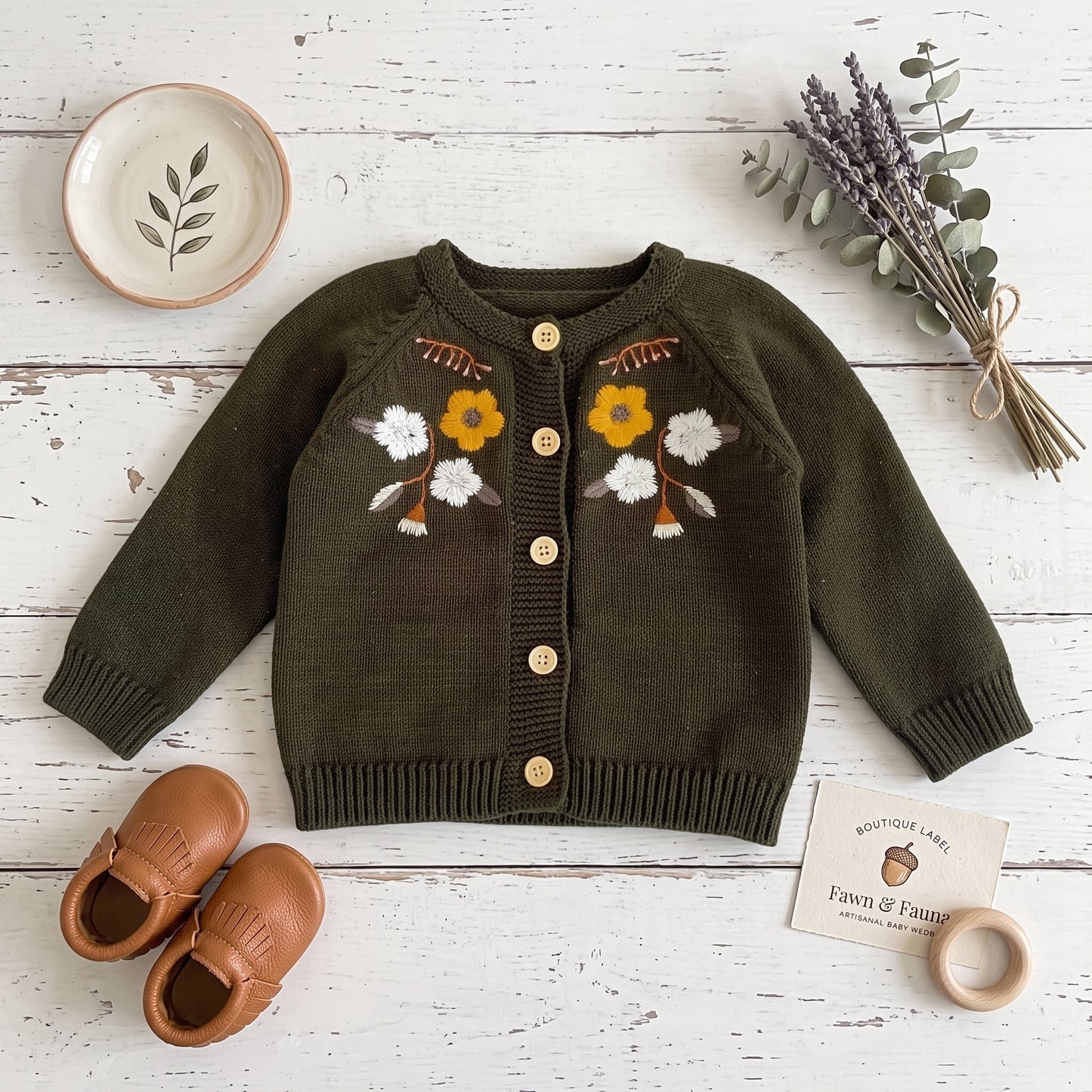 Flat lay view of olive green floral embroidered baby sweater paired with leather shoes and dried lavender.