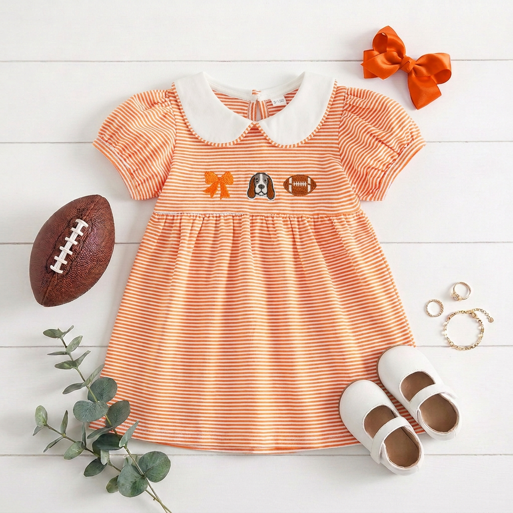 Flat lay of the toddler girls orange and white striped game day dress featuring hound dog and football embroidery, styled on a white wooden surface with white toddler shoes, an orange hair bow, a football, and greenery.