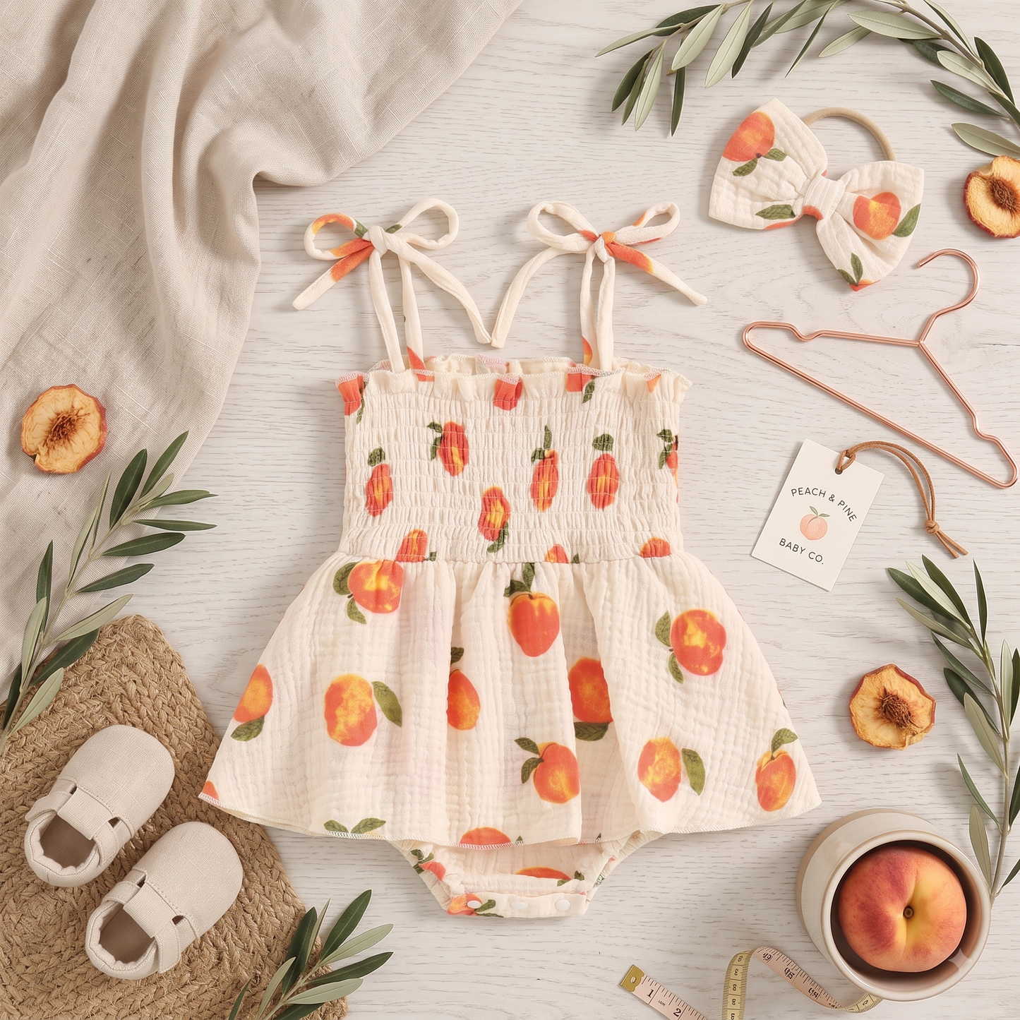 Flat lay of the baby girl peach print romper dress and headband styled with baby shoes, a hanger, and greenery.