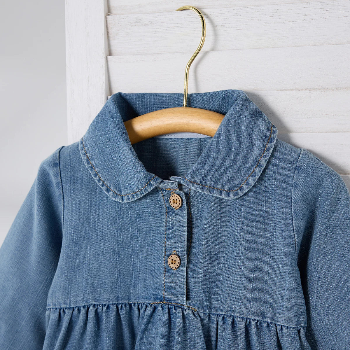 Close-up detail of the Peter Pan collar and wooden-style front buttons on a girls' blue denim jumpsuit on a hanger.