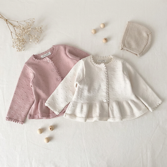 Two baby girl cotton knit ruffle cardigans in pink and ivory, laid flat on a light background next to a baby bonnet and small wooden blocks.
