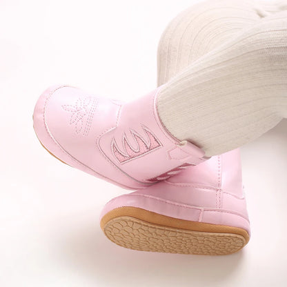 A baby wearing pink western cowboy booties with white tights, legs crossed to show the toe embroidery and metallic details.