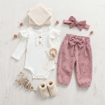 Styled flat lay of baby girl fall clothing set including a white ruffled romper, pink floral corduroy pants, headband, and baby booties.
