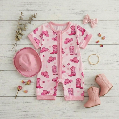 Flat lay of a pink baby girl western romper styled with a miniature pink cowgirl hat, pink boots, and dried flowers on a white wooden surface.