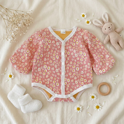 Styled flat lay of the pink daisy floral baby romper on a textured blanket, paired with baby socks, a wooden teething ring, and a plush bunny toy.