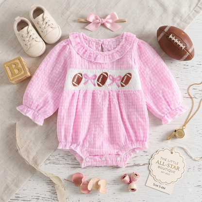 Styled flat lay of a baby girl pink smocked football bodysuit paired with baby shoes, a hair bow, and a mini football.