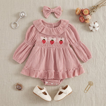Flat lay of a pink strawberry baby romper outfit styled with a matching headband, white baby shoes, and a silver rattle on a neutral background.